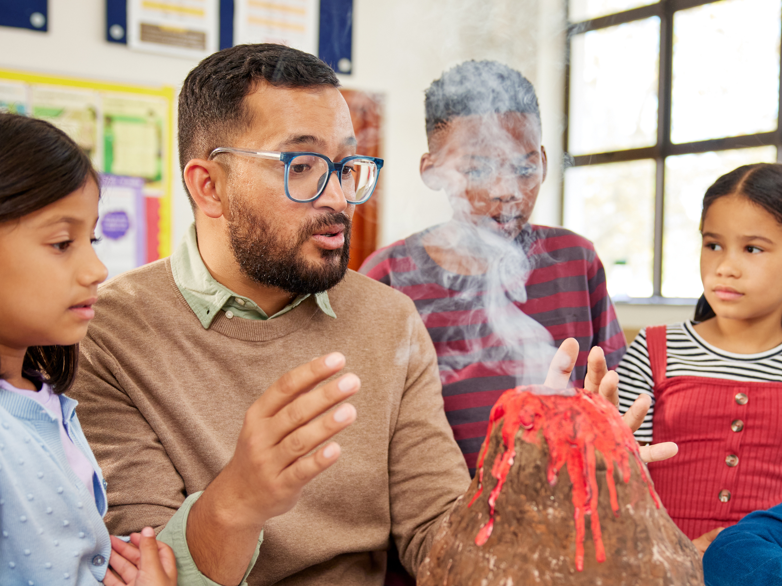 A teacher wearing glasses demonstrates a science experiment with a small erupting volcano model to a group of three students in a classroom. The students, two girls and a boy, watch attentively as the teacher explains the process. White smoke rises from the volcano, adding a sense of excitement to the scene. Colorful educational posters are visible in the background, contributing to the learning environment.