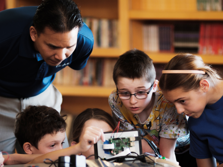 "Teacher guiding a group of engaged students as they collaborate on a hands-on STEM project involving circuitry and robotics in a library setting."