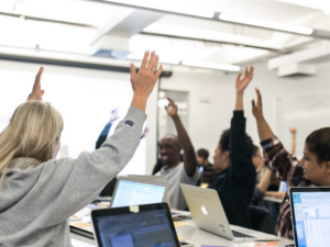 A group of individuals seated at desks in a bright classroom, raising their hands in unison during an engaging activity. Open laptops and notebooks are visible, emphasizing a collaborative and interactive learning environment.