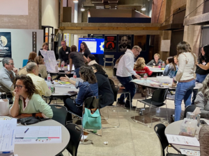 A collaborative workshop setting with participants seated at round tables, engaging in discussions and working on group projects. The room has an industrial, modern design with posters and a presentation screen in the background.